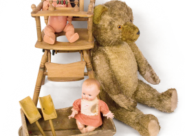 Plush teddy bear with a doll of German origin, sitting in a chair that converts to a stroller with wheels. This dates back to the 1920s and it is accompanied by a wooden wagon with a doll from the early 20th century